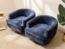 Load image into Gallery viewer, Two matching barrel-style swivel armchairs upholstered in deep navy blue velvet, placed on a beige carpeted floor against a white brick wall with a potted plant nearby.
