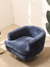 Load image into Gallery viewer, A modern barrel-style swivel armchair upholstered in textured navy blue velvet, set on a round metallic base in a bright room with a white brick wall and beige carpet.
