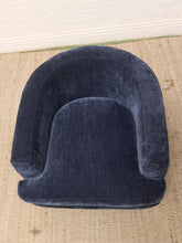 Load image into Gallery viewer, A curved, low-profile accent chair upholstered in dark navy blue velvet, placed on a textured beige woven rug against a white brick wall.
