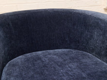Load image into Gallery viewer, Close-up of a curved upholstered chair in dark navy blue velvet fabric, showing the textured fabric and stitching against a light brick wall background.
