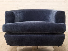 Load image into Gallery viewer, A modern barrel-style swivel armchair upholstered in dark navy blue velvet, featuring a circular brushed metal base, set against a white brick wall background.

