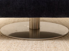 Load image into Gallery viewer, Close-up of a round brushed metal pedestal base supporting a dark upholstered furniture piece on a woven beige rug.

