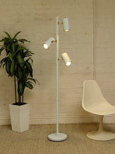 Load image into Gallery viewer, A modern white floor lamp with three adjustable cylindrical light heads stands in a room corner against a white brick wall, next to a potted green plant and a white molded chair.
