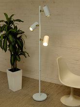 Load image into Gallery viewer, A modern white floor lamp with three adjustable cylindrical light heads and brass-colored joints, standing in a room with a white brick wall, a potted green plant, and a white molded chair.
