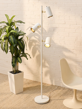 Load image into Gallery viewer, A modern three-light floor lamp with white cylindrical shades and gold-toned metal accents, standing in a room with a white brick wall, a potted green plant, and a cream-colored chair.
