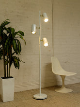 Load image into Gallery viewer, A modern white floor lamp with three adjustable cylindrical lights stands in a room corner against a white brick wall, next to a white molded chair and a potted green plant.

