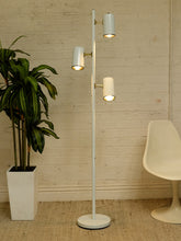 Load image into Gallery viewer, A modern three-light floor lamp with white cylindrical shades and gold-toned metal accents, standing in a room with a white brick wall, a potted plant, and a white chair.
