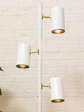 Load image into Gallery viewer, A modern three-light floor lamp with a white pole and cylindrical shades, featuring gold-toned metal accents and a white brick wall background.
