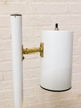Load image into Gallery viewer, A mid-century modern floor lamp featuring a white cylindrical shade and pole with a polished brass wall mount, set against a white brick wall background.

