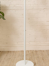 Load image into Gallery viewer, A minimalist white floor lamp with a circular base stands on a woven jute rug against a white brick wall, with a green plant leaf visible in the upper left corner.
