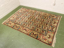 Load image into Gallery viewer, A rectangular Persian-style area rug with a beige background and intricate red, blue, and green floral patterns, laid out on a green floor against white brick walls.
