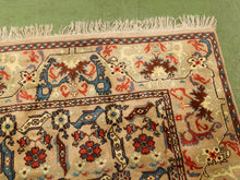 Load image into Gallery viewer, A close-up of a traditional hand-knotted area rug with a beige background, featuring red, blue, and brown floral and geometric motifs, laid out on a green floor.
