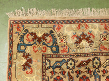Load image into Gallery viewer, A close-up of a traditional patterned rug with cream, blue, red, and brown motifs on a beige field, showing part of the woven design and fringe at the top against a green floor.
