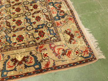 Load image into Gallery viewer, A close-up of a traditional patterned rug corner, showing cream, red, blue, and brown floral motifs with fringe, laid on a green floor.
