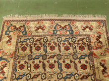 Load image into Gallery viewer, A traditional patterned area rug with cream, red, blue, and brown motifs, laid flat on a green floor. The rug features a central field with repeating floral and geometric designs, a decorative border, and fringed edges.
