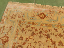 Load image into Gallery viewer, A close-up of a vintage-style patterned rug with a beige base, terracotta brown motifs, and light blue accents, featuring a fringed edge and laid out on a green floor.

