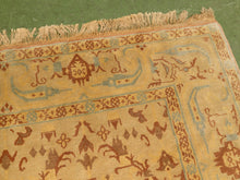 Load image into Gallery viewer, A close-up of a traditional patterned rug with a warm beige base, brown motifs, and light blue accents, featuring a fringed edge on a green surface.
