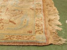 Load image into Gallery viewer, A close-up of an antique-style area rug corner, showing a beige and light blue floral pattern with rust-brown borders and fringed edges, laid on a green floor.
