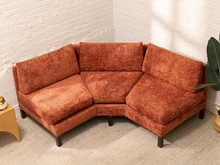 Load image into Gallery viewer, An L-shaped corner sectional sofa upholstered in textured rust-orange fabric, shown in a styled room corner with a white brick wall, beige carpet, a small wooden side table, and a potted plant.
