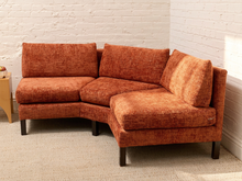 Load image into Gallery viewer, An L-shaped sectional sofa upholstered in textured rust-orange fabric, shown in a room corner with a white brick wall and light beige carpet.
