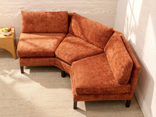 Load image into Gallery viewer, An L-shaped sectional sofa upholstered in textured rust-orange fabric, positioned in a room with a white brick wall and a light-colored rug.
