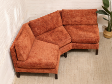 Load image into Gallery viewer, An L-shaped sectional sofa upholstered in textured rust-orange fabric, positioned in a room corner with a white brick wall, light-colored rug, and a potted plant.

