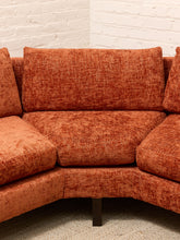 Load image into Gallery viewer, A close-up of a curved upholstered sofa in a textured rust-orange fabric, showing the seat cushion, back cushions, and part of the armrests, set against a white brick wall and light carpet.
