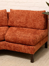 Load image into Gallery viewer, A close-up of a modern sectional sofa upholstered in a textured rust-orange bouclé fabric, shown with dark wooden legs against a white brick wall and light rug.
