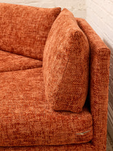 Load image into Gallery viewer, Close-up of an orange-brown textured bouclé fabric sofa with a matching back cushion, showing the woven upholstery detail against a white brick wall background.
