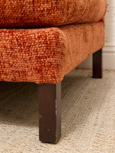 Load image into Gallery viewer, Close-up of an upholstered ottoman or stool corner in textured rust-orange fabric with dark wooden legs, shown on a woven rug indoors.
