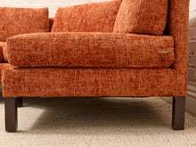 Load image into Gallery viewer, Close-up of an orange textured fabric armchair with dark wooden legs, positioned on a light beige woven rug against a white brick wall.
