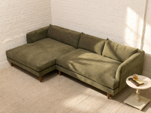 Load image into Gallery viewer, An olive green textured fabric sectional sofa with a chaise lounge, positioned in a bright room with white brick walls and a light-colored rug, next to a small white side table.
