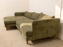 Load image into Gallery viewer, An olive green sectional sofa with a chaise lounge on the left, upholstered in a textured waffle-weave fabric, shown in a room with a white brick wall and beige carpet.
