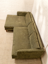 Load image into Gallery viewer, An olive green sectional sofa with a textured, woven basketweave pattern, positioned in a room with a white brick wall and a light beige rug.
