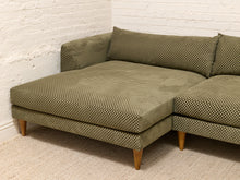 Load image into Gallery viewer, An olive green sectional sofa with a textured geometric pattern, featuring a chaise lounge on the left and tapered wooden legs, set against a white brick wall and light beige carpet.
