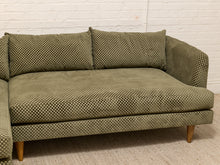 Load image into Gallery viewer, A mid-century modern corner sofa upholstered in olive green fabric with a small-scale geometric pattern, featuring light wood legs, set against a white brick wall background.
