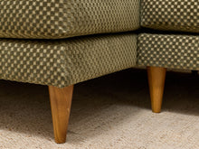 Load image into Gallery viewer, Close-up of the corner of a mid-century modern sofa upholstered in a textured olive green and beige checkerboard pattern, supported by tapered wooden legs on a beige carpet.
