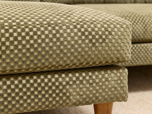Load image into Gallery viewer, Close-up of an upholstered ottoman or sofa cushion with a textured checkerboard pattern in beige and olive green, showing part of the wooden leg and carpet.
