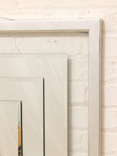 Load image into Gallery viewer, A close-up of a large white rectangular frame leaning against a white brick wall, with two smaller matching frames partially visible behind it.
