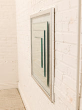 Load image into Gallery viewer, A rectangular wall mirror with a thin silver frame hangs on a white painted brick wall. The mirror reflects a dark green interior space, and a light beige woven rug is visible in the lower-left corner.

