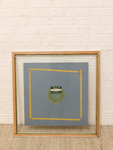 Load image into Gallery viewer, A square framed art print with a light wood frame and clear glass, leaning against a white brick wall on a beige carpet. The artwork features a gray background with a yellow geometric square border and a green circular center containing a small label.
