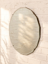 Load image into Gallery viewer, An oval wall mirror with a dark metal frame hanging on a white painted brick wall, with soft leaf shadows cast across the reflective surface.
