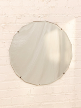 Load image into Gallery viewer, A large round wall mirror with a thin dark metal frame, mounted on a white painted brick wall with soft light casting a shadow across the reflective surface.
