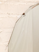 Load image into Gallery viewer, A close-up of a large, off-white oval mirror with a thin metallic frame, set against a textured, off-white brick wall.
