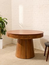 Load image into Gallery viewer, A round wooden dining table with a fluted apron and tapered pedestal base, shown in a bright room with a white brick wall, beige carpet, a potted plant, and part of a chair visible.
