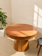 Load image into Gallery viewer, A round wooden dining table with a fluted rim and tapered pedestal base, styled in a bright room with a white brick wall, beige carpet, a potted plant, and part of a chair visible.
