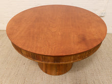 Load image into Gallery viewer, A round wooden coffee table with a warm reddish-brown finish, featuring a fluted pedestal base and a matching fluted rim, set on a textured beige rug against a white brick wall.
