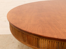Load image into Gallery viewer, Close-up of a round wooden table with a warm medium-brown finish, showing the tabletop and fluted lower edge against a light off-white brick wall and beige floor.

