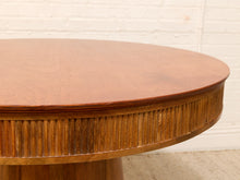Load image into Gallery viewer, Close-up of a round wooden dining table with a warm medium-brown finish, featuring a fluted pedestal base and a smooth oval tabletop, set against a white brick wall.
