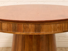 Load image into Gallery viewer, Close-up of a round wooden table with a warm medium-brown finish, featuring a fluted decorative apron and a thick pedestal base, set against a white brick wall.
